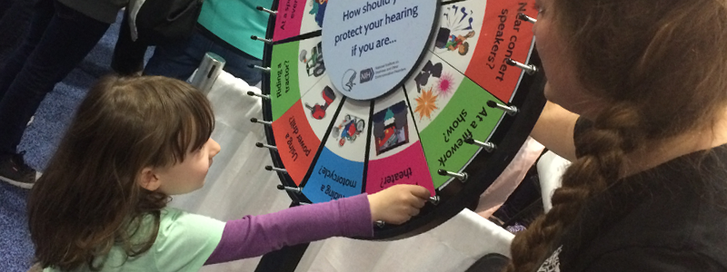 Using the Noisy Planet Q/A spinning wheel, a volunteer teaches a visitor about healthy hearing habits for common noises. (National Institute on Deafness and Other Communication Disorders)
