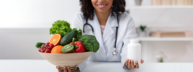Smiling doctor holding a bowl of fruits and vegetables in one hand and bottle of medicine in the other
